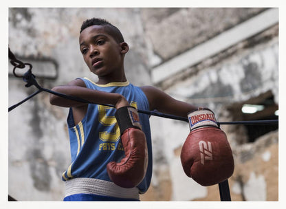 A young Black boy in a blue jersey and red boxing gloves leans pensively against the ropes of a boxing ring, looking away with a thoughtful expression against a blurred, weathered background. Wall Art