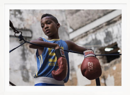 A young Black boy in a blue jersey and red boxing gloves leans pensively against the ropes of a boxing ring, looking away with a thoughtful expression against a blurred, weathered background. Wall Art