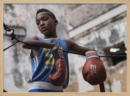 A young Black boy in a blue jersey and red boxing gloves leans pensively against the ropes of a boxing ring, looking away with a thoughtful expression against a blurred, weathered background. Wall Art