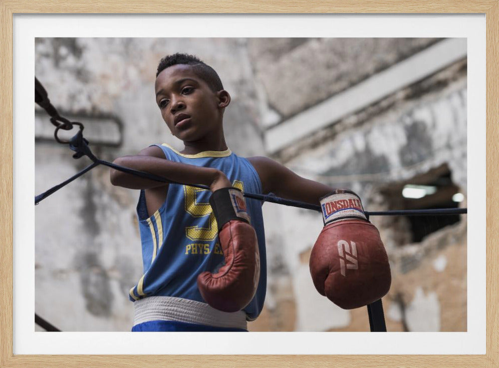A young Black boy in a blue jersey and red boxing gloves leans pensively against the ropes of a boxing ring, looking away with a thoughtful expression against a blurred, weathered background. Wall Art