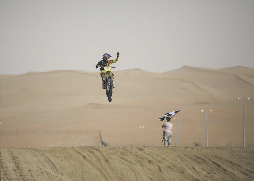 A motocross rider on a yellow dirt bike is captured mid-air during a jump in a desert landscape, flashing a peace sign with their left hand. In the background, rolling sand dunes stretch under a hazy sky, and to the right, a person on a hill waves a black and white checkered flag. Print