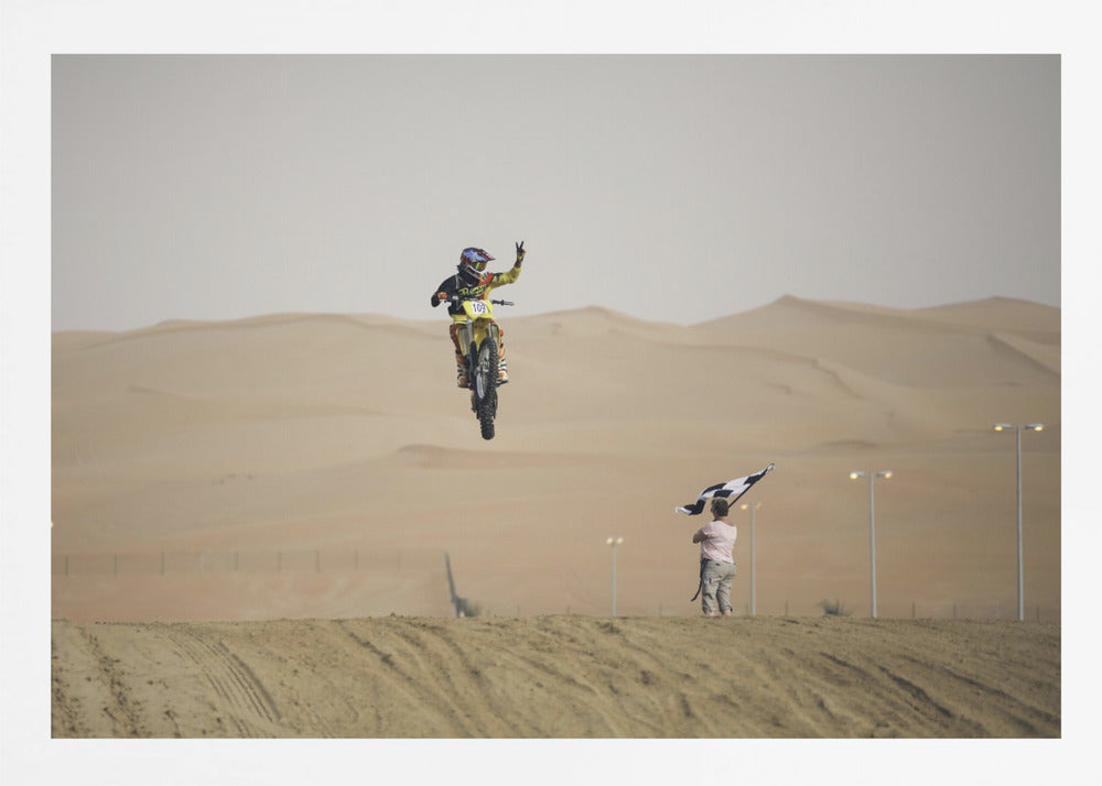 A motocross rider on a yellow dirt bike is captured mid-air during a jump in a desert landscape, flashing a peace sign with their left hand. In the background, rolling sand dunes stretch under a hazy sky, and to the right, a person on a hill waves a black and white checkered flag. Print