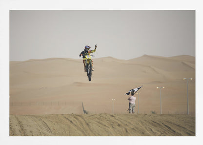 A motocross rider on a yellow dirt bike is captured mid-air during a jump in a desert landscape, flashing a peace sign with their left hand. In the background, rolling sand dunes stretch under a hazy sky, and to the right, a person on a hill waves a black and white checkered flag. Print