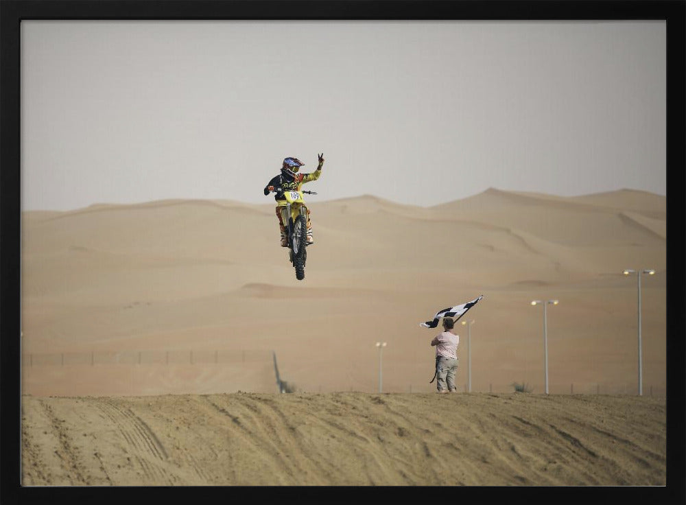 A motocross rider on a yellow dirt bike is captured mid-air during a jump in a desert landscape, flashing a peace sign with their left hand. In the background, rolling sand dunes stretch under a hazy sky, and to the right, a person on a hill waves a black and white checkered flag. Print