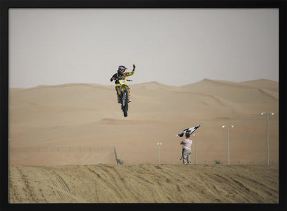 A motocross rider on a yellow dirt bike is captured mid-air during a jump in a desert landscape, flashing a peace sign with their left hand. In the background, rolling sand dunes stretch under a hazy sky, and to the right, a person on a hill waves a black and white checkered flag. Print