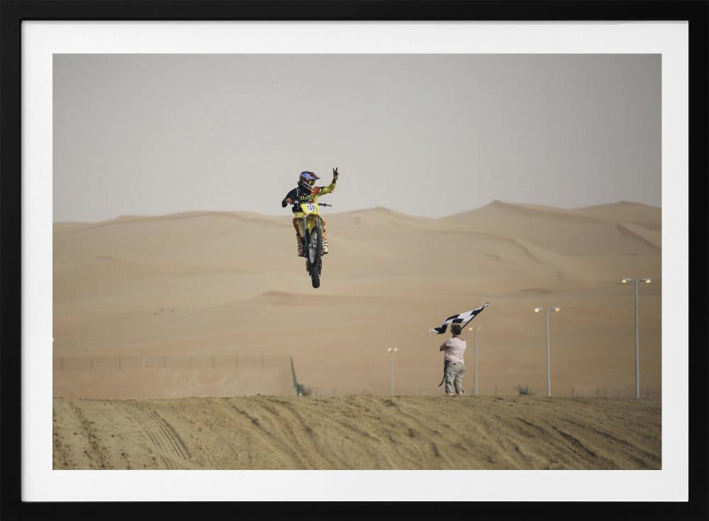 A motocross rider on a yellow dirt bike is captured mid-air during a jump in a desert landscape, flashing a peace sign with their left hand. In the background, rolling sand dunes stretch under a hazy sky, and to the right, a person on a hill waves a black and white checkered flag. Print