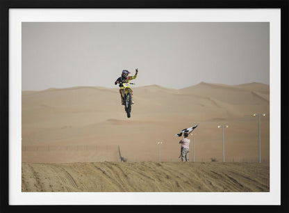 A motocross rider on a yellow dirt bike is captured mid-air during a jump in a desert landscape, flashing a peace sign with their left hand. In the background, rolling sand dunes stretch under a hazy sky, and to the right, a person on a hill waves a black and white checkered flag. Print