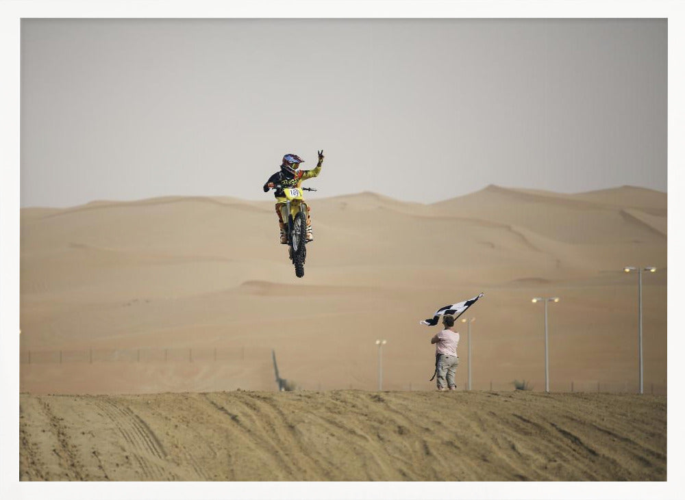 A motocross rider on a yellow dirt bike is captured mid-air during a jump in a desert landscape, flashing a peace sign with their left hand. In the background, rolling sand dunes stretch under a hazy sky, and to the right, a person on a hill waves a black and white checkered flag. Print