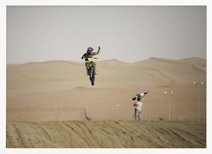 A motocross rider on a yellow dirt bike is captured mid-air during a jump in a desert landscape, flashing a peace sign with their left hand. In the background, rolling sand dunes stretch under a hazy sky, and to the right, a person on a hill waves a black and white checkered flag. Print
