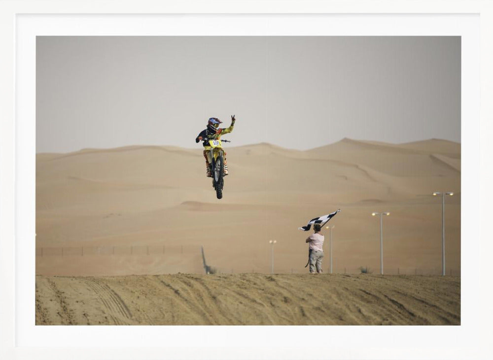 A motocross rider on a yellow dirt bike is captured mid-air during a jump in a desert landscape, flashing a peace sign with their left hand. In the background, rolling sand dunes stretch under a hazy sky, and to the right, a person on a hill waves a black and white checkered flag. Print