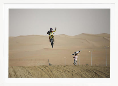 A motocross rider on a yellow dirt bike is captured mid-air during a jump in a desert landscape, flashing a peace sign with their left hand. In the background, rolling sand dunes stretch under a hazy sky, and to the right, a person on a hill waves a black and white checkered flag. Print