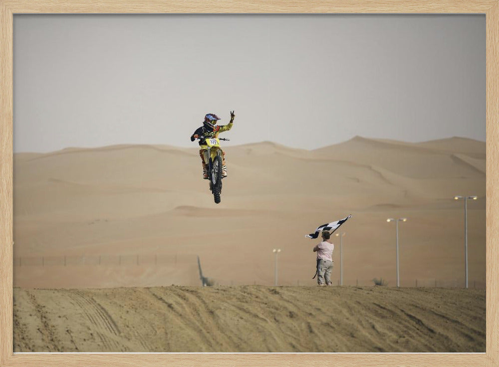 A motocross rider on a yellow dirt bike is captured mid-air during a jump in a desert landscape, flashing a peace sign with their left hand. In the background, rolling sand dunes stretch under a hazy sky, and to the right, a person on a hill waves a black and white checkered flag. Print