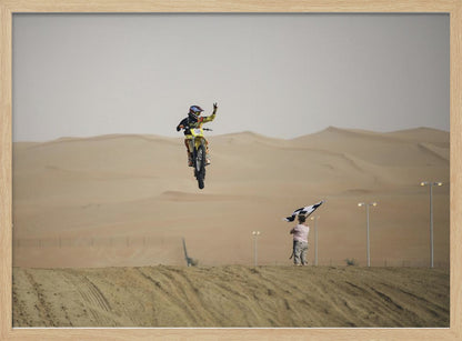A motocross rider on a yellow dirt bike is captured mid-air during a jump in a desert landscape, flashing a peace sign with their left hand. In the background, rolling sand dunes stretch under a hazy sky, and to the right, a person on a hill waves a black and white checkered flag. Print