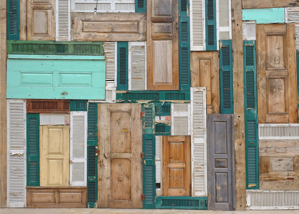 A framed art piece featuring a chaotic collage of various old, weathered wooden doors and window shutters arranged in a dense patchwork, showcasing a rustic texture and colors like natural wood, teal, green, and distressed white. Wall Art