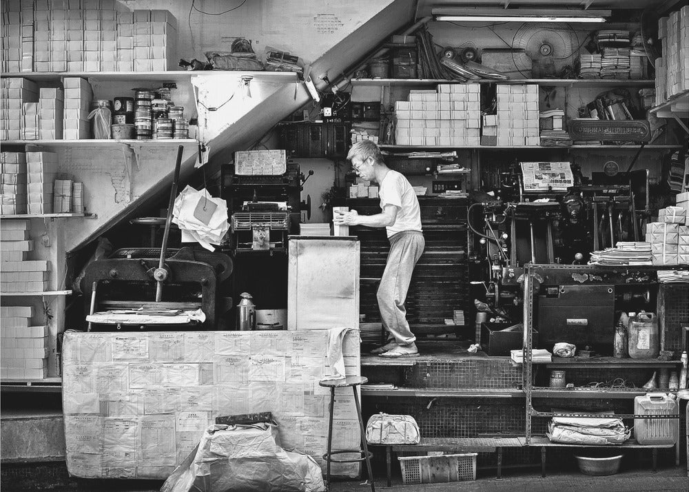 A vintage art print depicting a small, old-fashioned printing shop interior, showcasing antique machinery and a nostalgic atmosphere. Artwork