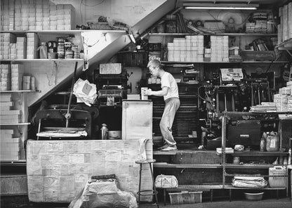 A vintage art print depicting a small, old-fashioned printing shop interior, showcasing antique machinery and a nostalgic atmosphere. Artwork