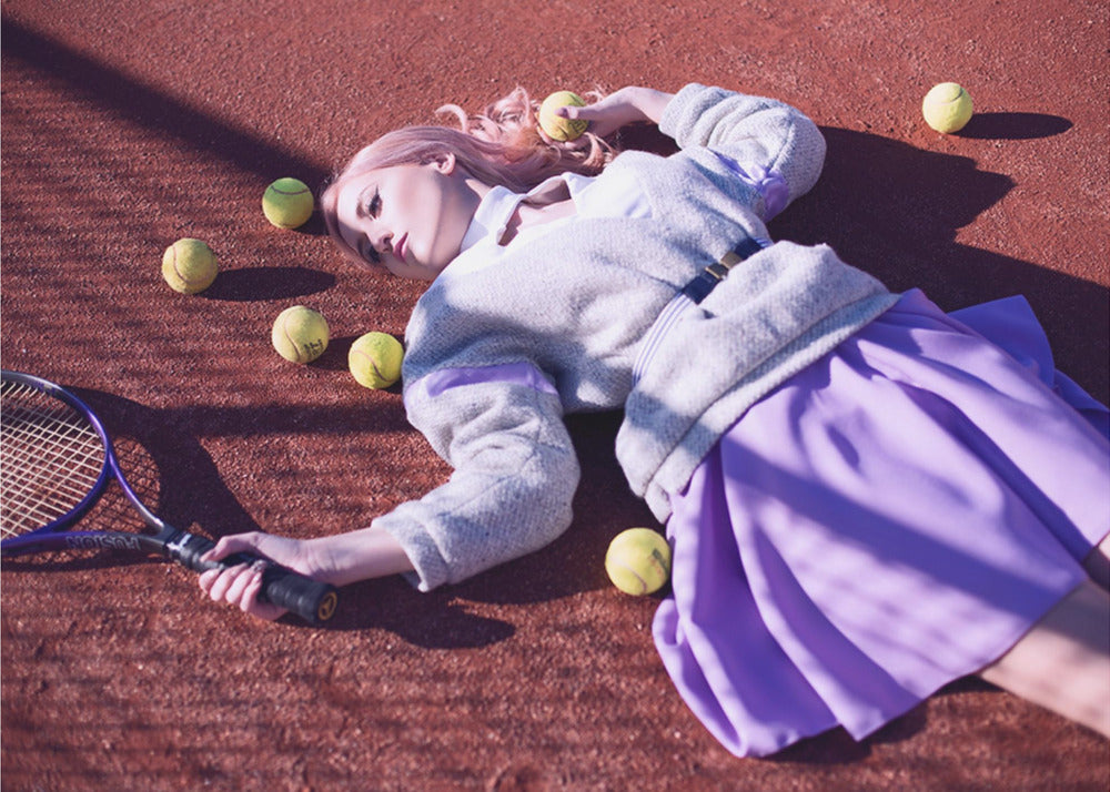 A high-angle shot of a young woman with pink hair lying on a red clay tennis court, surrounded by yellow tennis balls. She wears a grey sweater, a white collared shirt, and a purple pleated skirt, and holds a tennis racket in one hand. The scene is framed with a silver border. Wall Art