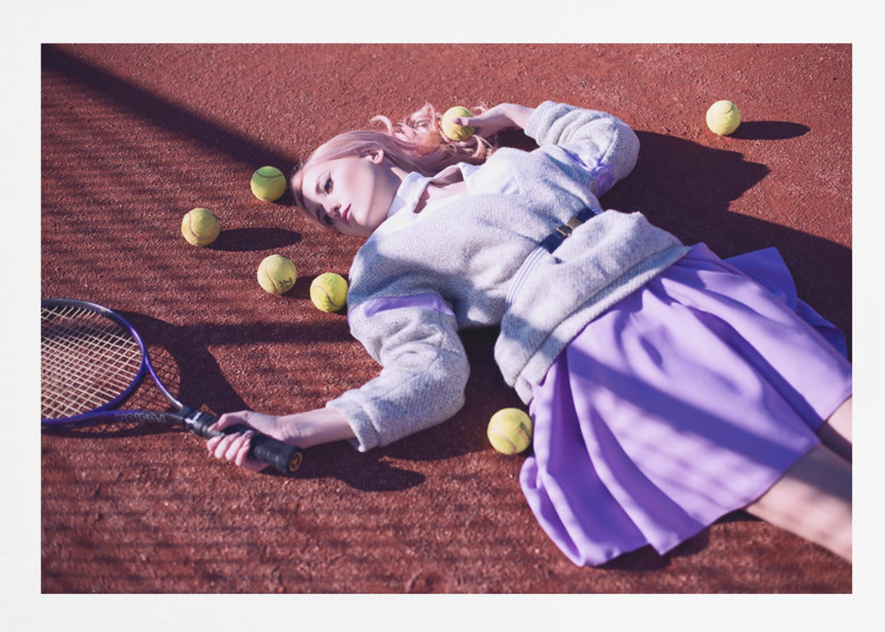 A high-angle shot of a young woman with pink hair lying on a red clay tennis court, surrounded by yellow tennis balls. She wears a grey sweater, a white collared shirt, and a purple pleated skirt, and holds a tennis racket in one hand. The scene is framed with a silver border. Wall Art