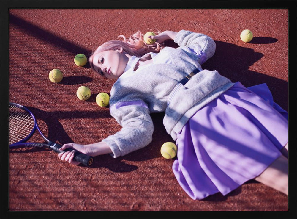 A high-angle shot of a young woman with pink hair lying on a red clay tennis court, surrounded by yellow tennis balls. She wears a grey sweater, a white collared shirt, and a purple pleated skirt, and holds a tennis racket in one hand. The scene is framed with a silver border. Wall Art