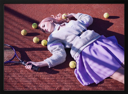 A high-angle shot of a young woman with pink hair lying on a red clay tennis court, surrounded by yellow tennis balls. She wears a grey sweater, a white collared shirt, and a purple pleated skirt, and holds a tennis racket in one hand. The scene is framed with a silver border. Wall Art
