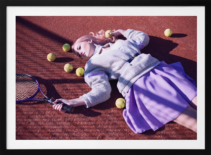 A high-angle shot of a young woman with pink hair lying on a red clay tennis court, surrounded by yellow tennis balls. She wears a grey sweater, a white collared shirt, and a purple pleated skirt, and holds a tennis racket in one hand. The scene is framed with a silver border. Wall Art