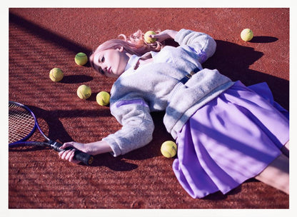 A high-angle shot of a young woman with pink hair lying on a red clay tennis court, surrounded by yellow tennis balls. She wears a grey sweater, a white collared shirt, and a purple pleated skirt, and holds a tennis racket in one hand. The scene is framed with a silver border. Wall Art