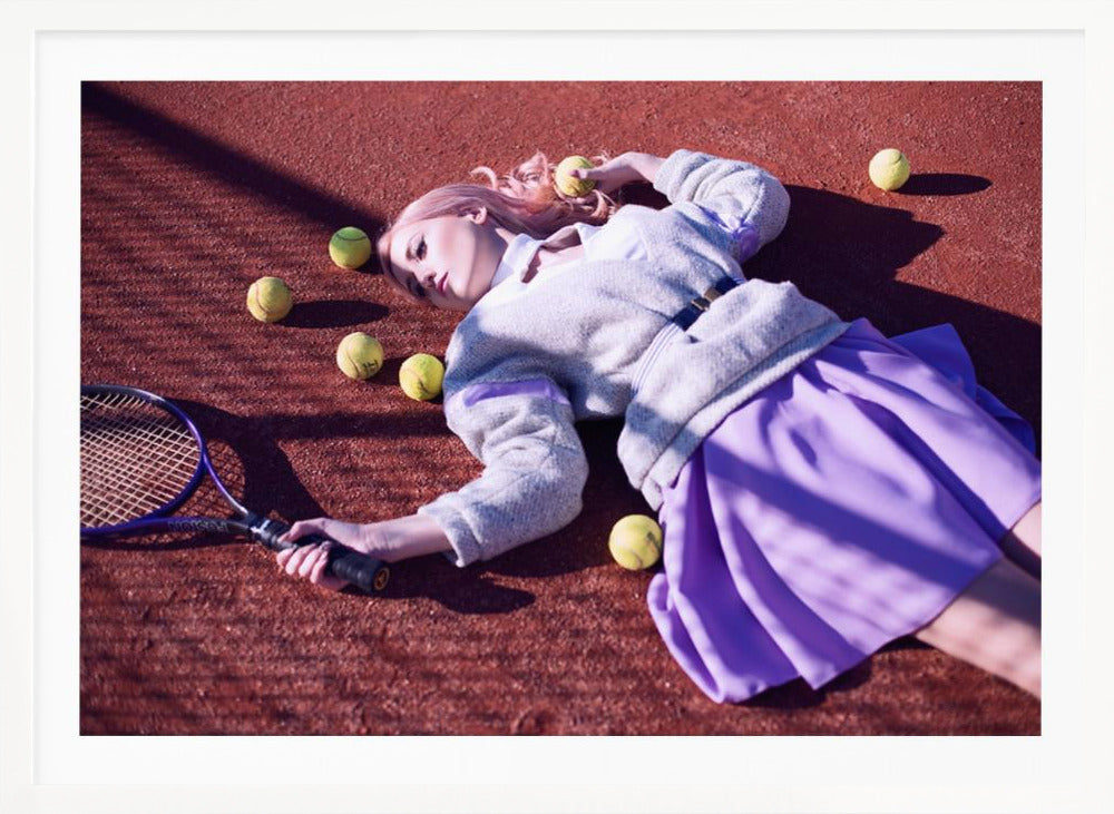 A high-angle shot of a young woman with pink hair lying on a red clay tennis court, surrounded by yellow tennis balls. She wears a grey sweater, a white collared shirt, and a purple pleated skirt, and holds a tennis racket in one hand. The scene is framed with a silver border. Wall Art