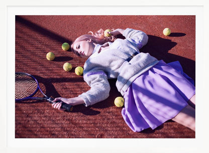 A high-angle shot of a young woman with pink hair lying on a red clay tennis court, surrounded by yellow tennis balls. She wears a grey sweater, a white collared shirt, and a purple pleated skirt, and holds a tennis racket in one hand. The scene is framed with a silver border. Wall Art