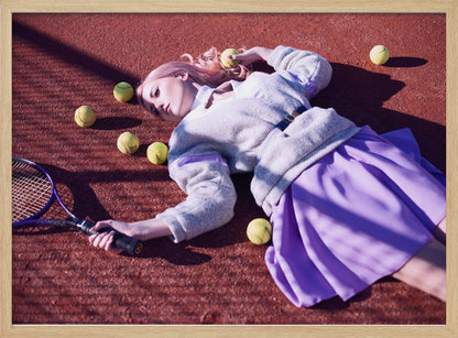 A high-angle shot of a young woman with pink hair lying on a red clay tennis court, surrounded by yellow tennis balls. She wears a grey sweater, a white collared shirt, and a purple pleated skirt, and holds a tennis racket in one hand. The scene is framed with a silver border. Wall Art