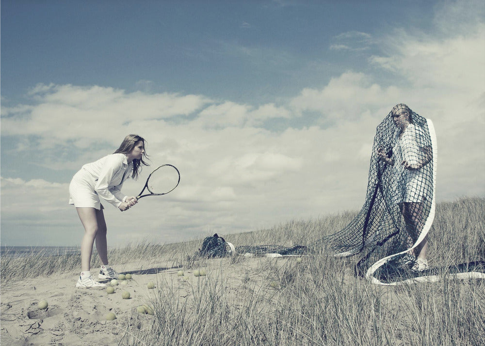 A surreal photograph of two women on a grassy sand dune. One woman is dressed in white tennis attire, bent over and ready to hit a ball with her racket. The other woman stands opposite her, entangled and draped in a large tennis net. The scene is under a cloudy blue sky and has a desaturated, cool-toned color palette, framed in silver. Decor