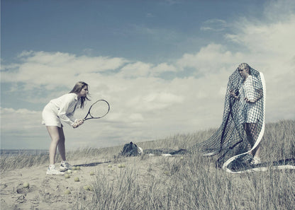 A surreal photograph of two women on a grassy sand dune. One woman is dressed in white tennis attire, bent over and ready to hit a ball with her racket. The other woman stands opposite her, entangled and draped in a large tennis net. The scene is under a cloudy blue sky and has a desaturated, cool-toned color palette, framed in silver. Decor