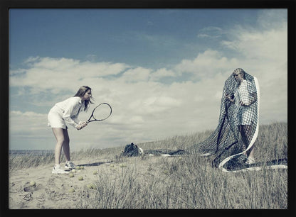 A surreal photograph of two women on a grassy sand dune. One woman is dressed in white tennis attire, bent over and ready to hit a ball with her racket. The other woman stands opposite her, entangled and draped in a large tennis net. The scene is under a cloudy blue sky and has a desaturated, cool-toned color palette, framed in silver. Decor