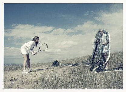 A surreal photograph of two women on a grassy sand dune. One woman is dressed in white tennis attire, bent over and ready to hit a ball with her racket. The other woman stands opposite her, entangled and draped in a large tennis net. The scene is under a cloudy blue sky and has a desaturated, cool-toned color palette, framed in silver. Decor