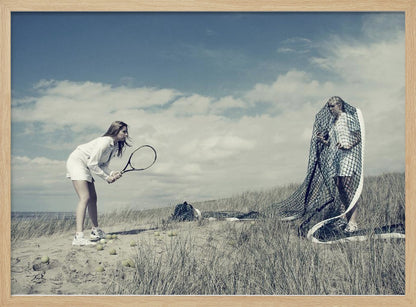 A surreal photograph of two women on a grassy sand dune. One woman is dressed in white tennis attire, bent over and ready to hit a ball with her racket. The other woman stands opposite her, entangled and draped in a large tennis net. The scene is under a cloudy blue sky and has a desaturated, cool-toned color palette, framed in silver. Decor