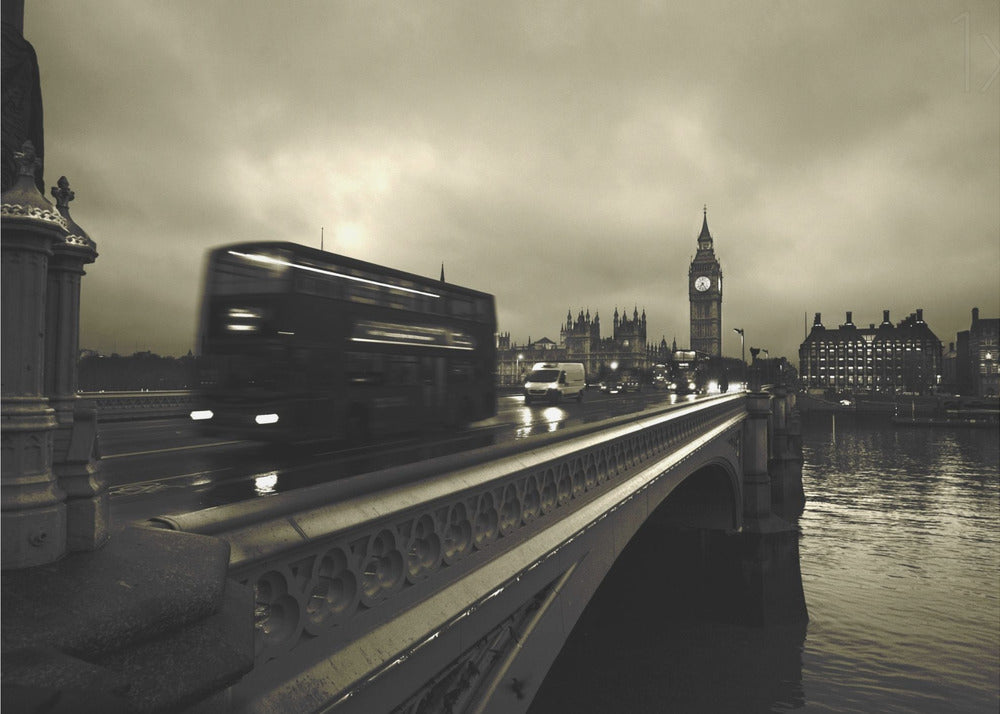 A framed, sepia-toned long-exposure photograph of London. A double-decker bus is shown in motion blur as it crosses Westminster Bridge. In the background, the Big Ben clock tower and the Houses of Parliament are visible against a cloudy sky, with lights reflecting on the wet road and the River Thames. Wall Art