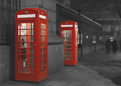 A selective color photograph of a city street at night, framed in silver. Two iconic red telephone booths are the only objects in color, standing out against the black and white background of the cobblestone sidewalk and iron fence. Artwork