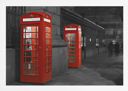 A selective color photograph of a city street at night, framed in silver. Two iconic red telephone booths are the only objects in color, standing out against the black and white background of the cobblestone sidewalk and iron fence. Artwork