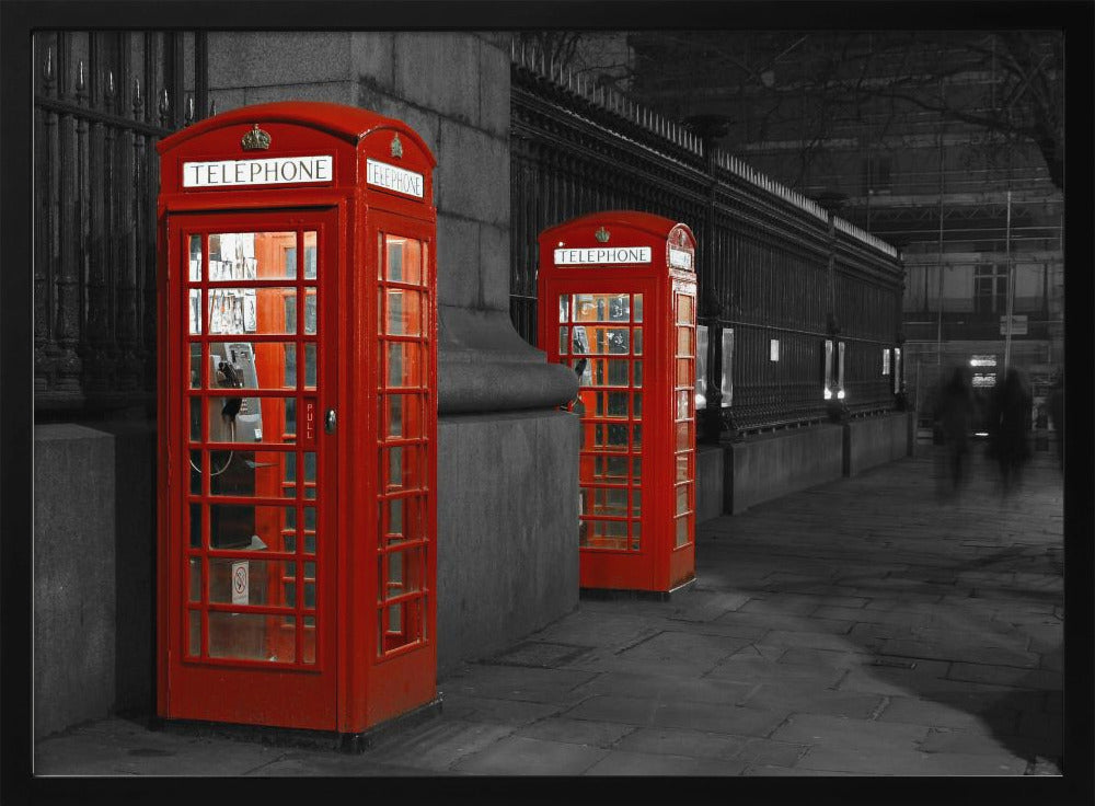 A selective color photograph of a city street at night, framed in silver. Two iconic red telephone booths are the only objects in color, standing out against the black and white background of the cobblestone sidewalk and iron fence. Artwork