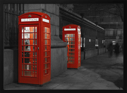 A selective color photograph of a city street at night, framed in silver. Two iconic red telephone booths are the only objects in color, standing out against the black and white background of the cobblestone sidewalk and iron fence. Artwork