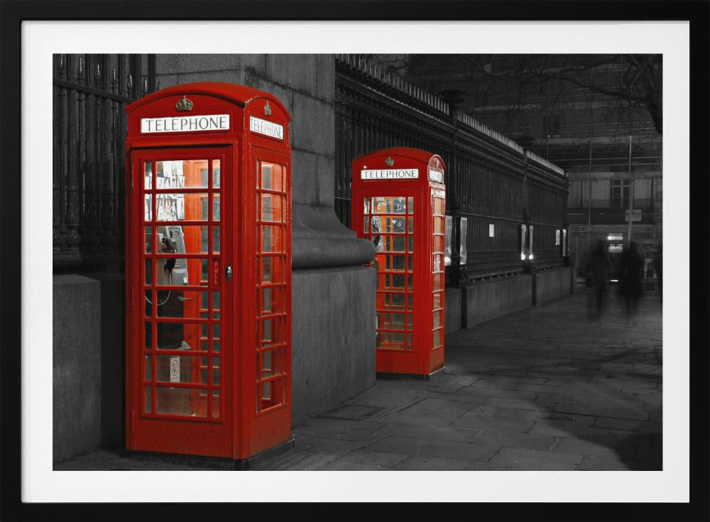 A selective color photograph of a city street at night, framed in silver. Two iconic red telephone booths are the only objects in color, standing out against the black and white background of the cobblestone sidewalk and iron fence. Artwork