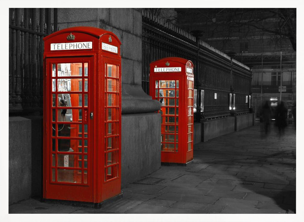 A selective color photograph of a city street at night, framed in silver. Two iconic red telephone booths are the only objects in color, standing out against the black and white background of the cobblestone sidewalk and iron fence. Artwork