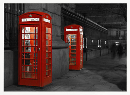A selective color photograph of a city street at night, framed in silver. Two iconic red telephone booths are the only objects in color, standing out against the black and white background of the cobblestone sidewalk and iron fence. Artwork