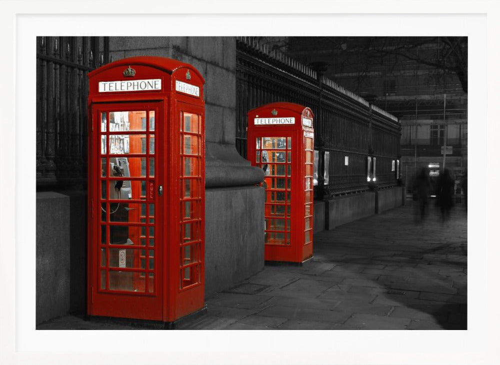 A selective color photograph of a city street at night, framed in silver. Two iconic red telephone booths are the only objects in color, standing out against the black and white background of the cobblestone sidewalk and iron fence. Artwork