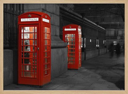 A selective color photograph of a city street at night, framed in silver. Two iconic red telephone booths are the only objects in color, standing out against the black and white background of the cobblestone sidewalk and iron fence. Artwork