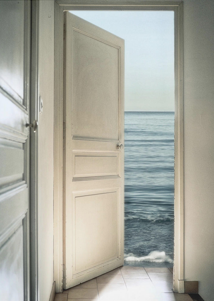 A surreal photograph showing a white interior door swung open, revealing not a landscape but the vast blue ocean, with waves gently washing over the tiled floor of the room. Decor