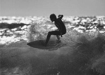 A black and white action shot of a surfer silhouetted against the sparkling ocean. The surfer is crouched low on their board, carving through a wave and creating a dramatic spray of water. The photo is enclosed in a silver frame. Print