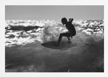 A black and white action shot of a surfer silhouetted against the sparkling ocean. The surfer is crouched low on their board, carving through a wave and creating a dramatic spray of water. The photo is enclosed in a silver frame. Print