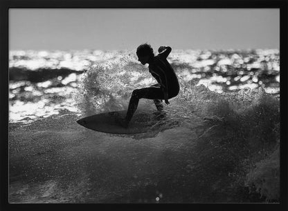 A black and white action shot of a surfer silhouetted against the sparkling ocean. The surfer is crouched low on their board, carving through a wave and creating a dramatic spray of water. The photo is enclosed in a silver frame. Print