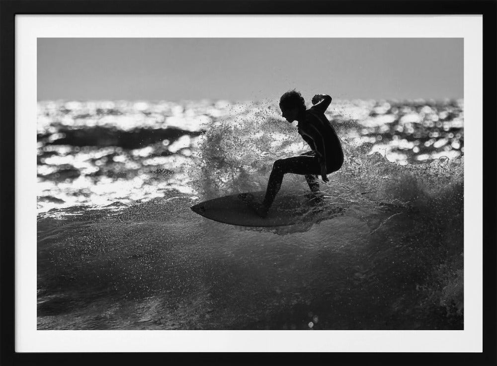 A black and white action shot of a surfer silhouetted against the sparkling ocean. The surfer is crouched low on their board, carving through a wave and creating a dramatic spray of water. The photo is enclosed in a silver frame. Print