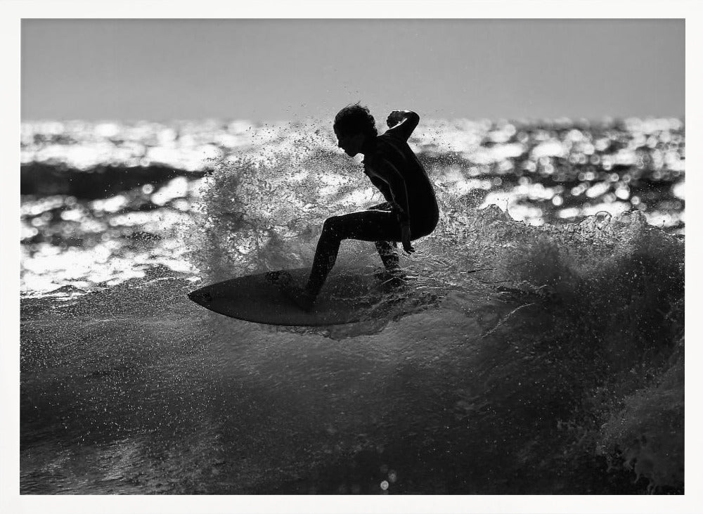 A black and white action shot of a surfer silhouetted against the sparkling ocean. The surfer is crouched low on their board, carving through a wave and creating a dramatic spray of water. The photo is enclosed in a silver frame. Print